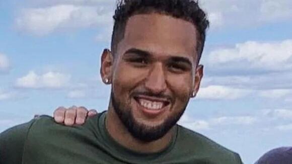 A young man with short curly hair and a beard smiles at the camera while wearing a green athletic shirt. The background features a bright blue sky with scattered clouds. His arm is around someone partially visible beside him.